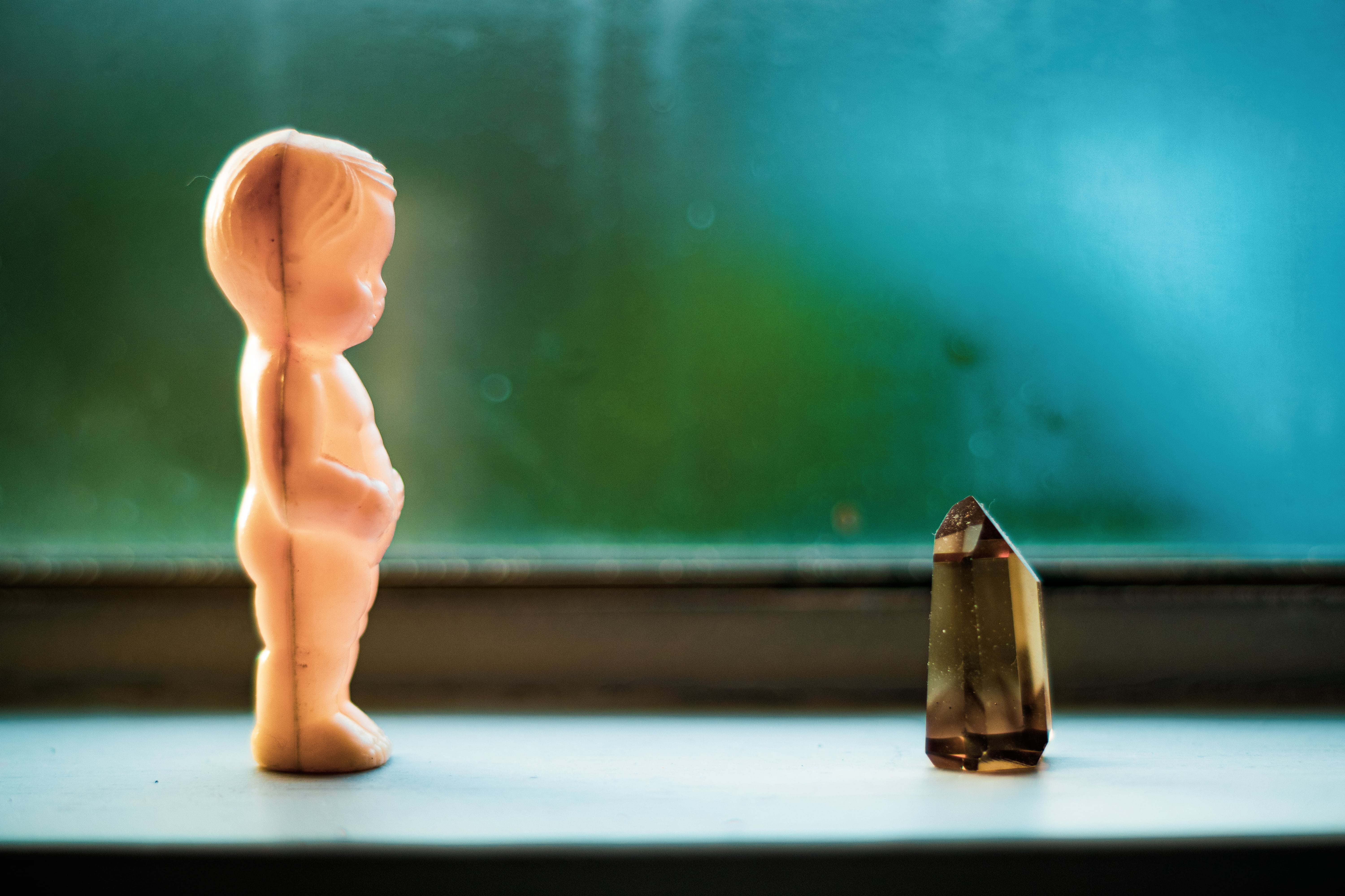 A photo of a small plastic baby standing on a window ledge facing a brown piece of quartz crystal.